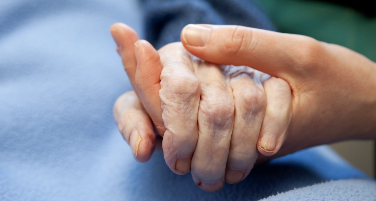 A caring hand holds that of an elderly patient with a blue blanket as background.