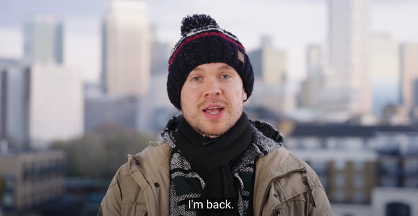 Gary Stevenson in a coat, scarf and hat on the roof of a building, saying "I'm back".