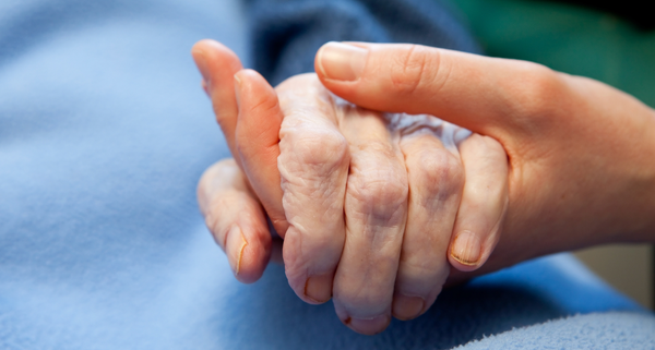 A caring hand holds that of an elderly patient with a blue blanket as background.